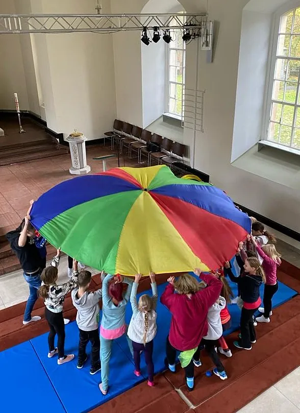 Kinder stehen in einer Kirche auf Turnmatten im Kreis und halten gemeinsam ein großes buntes Schwungtuch (Regenbogentuch) hoch, das wie ein Dach über ihnen gespannt ist.